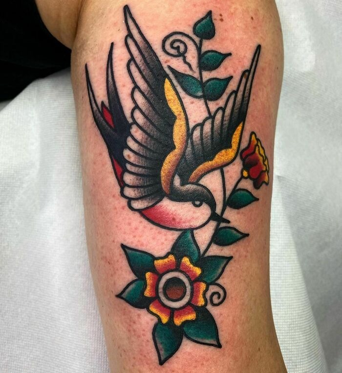 American traditional tattoo featuring a flying bird and vibrant flowers, showcasing classic bold colors and designs.
