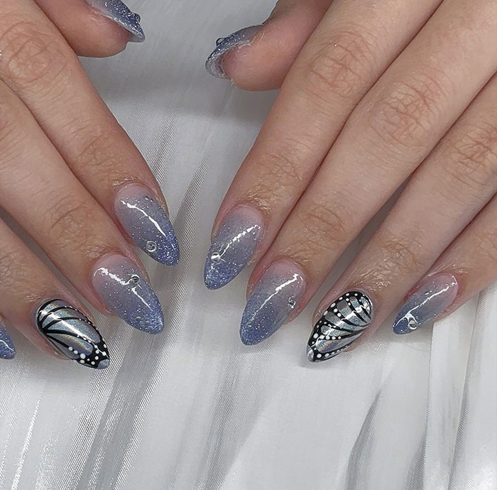Close-up of hands with trendy almond nails featuring blue glitter ombre and black silver butterfly designs.