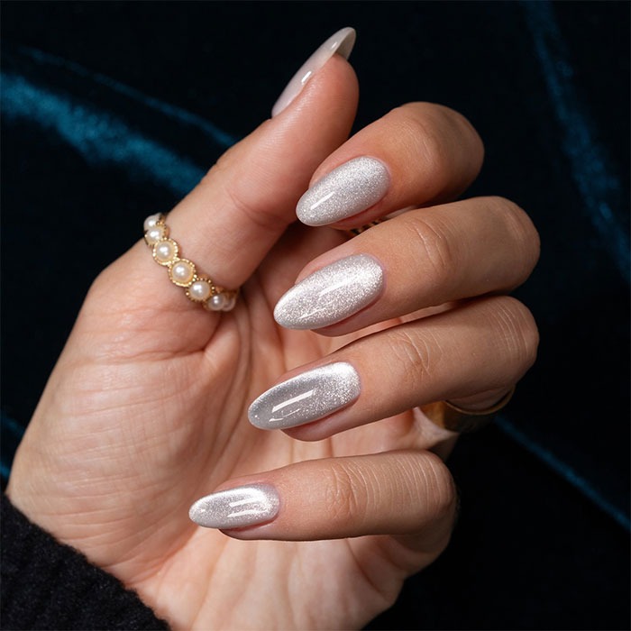 Hand with almond nails painted in shimmering silver polish, wearing pearl and gold rings against a dark background.