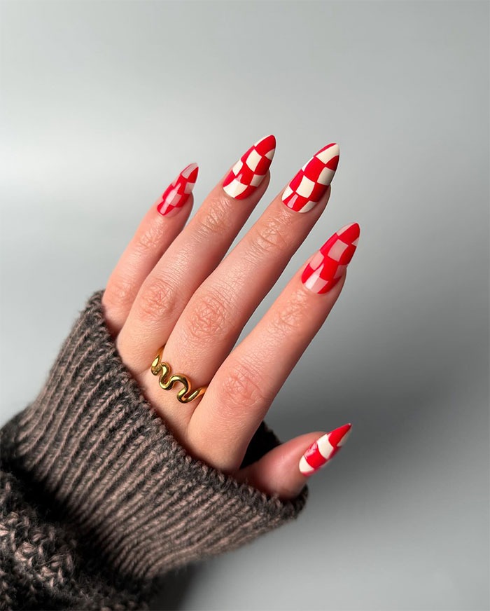 Hand with red and cream checkerboard almond nails, wearing a gold wavy ring and a brown knitted sweater sleeve.